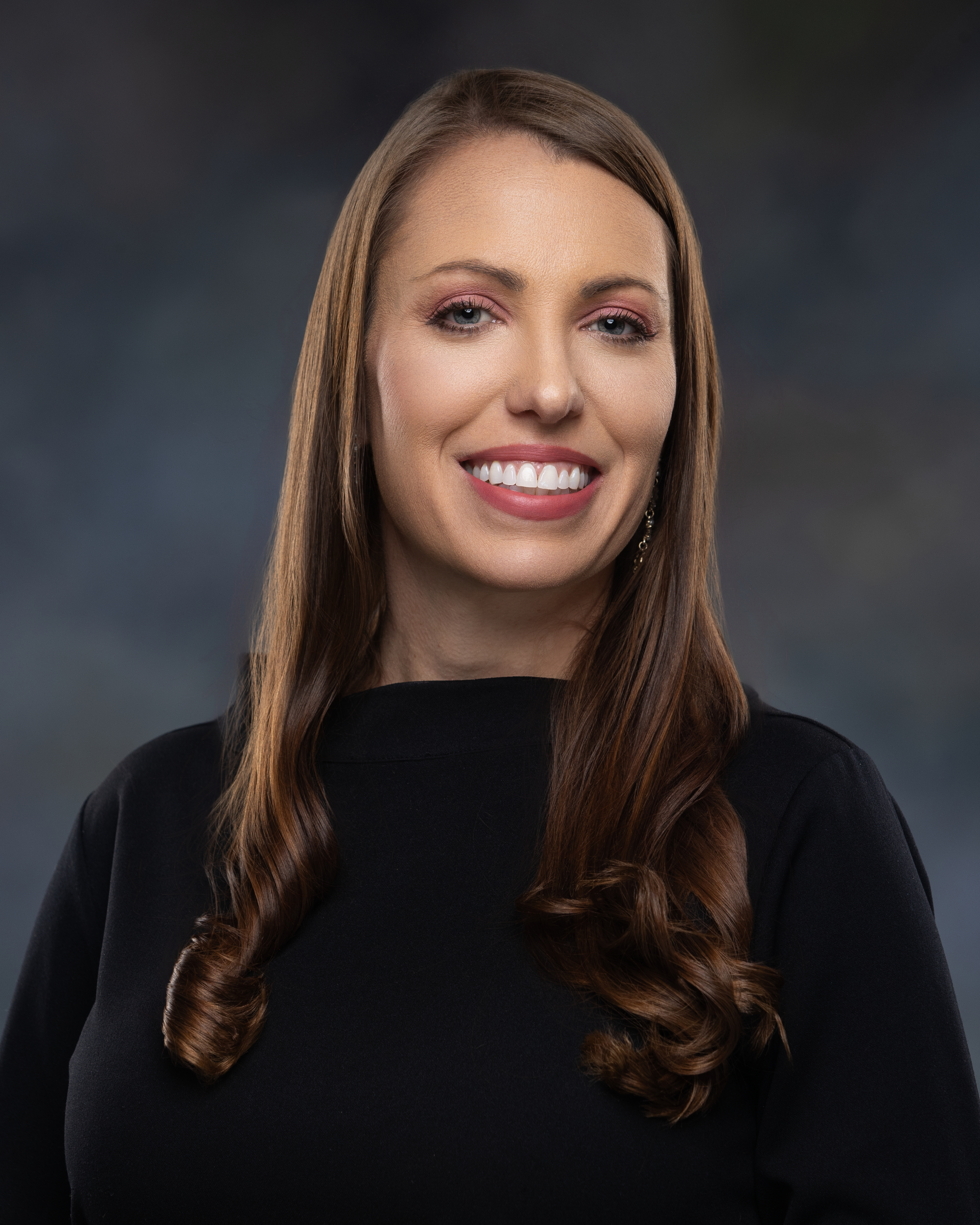 Woman with long brown hair wearing a black sweater smiles in front of a dark gray background